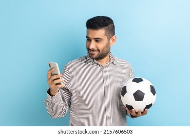 Portrait of smiling optimistic handsome bearded businessman standing with soccer ball and using mobile phone, wearing striped shirt. Indoor studio shot isolated on blue background. - Powered by Shutterstock