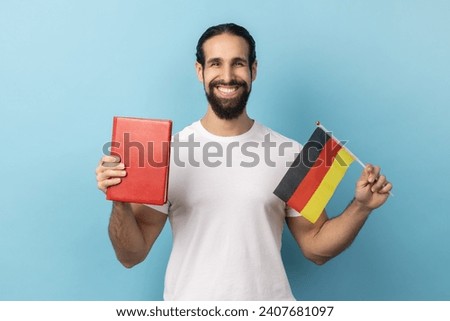 Portrait of smiling optimistic man with beard wearing white T-shirt holding book and Germany flag, education, learning german language. Indoor studio shot isolated on blue background.