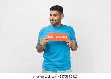 Portrait of smiling optimistic unshaven man in blue T- shirt standing holding card with subscribe inscription, looking at camera with happy expression. Indoor studio shot isolated on gray background. - Powered by Shutterstock