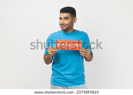 Portrait of smiling optimistic unshaven man in blue T- shirt standing holding card with subscribe inscription, looking at camera with happy expression. Indoor studio shot isolated on gray background.