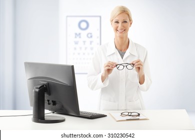 Portrait Of Smiling Optometrist Doctor Standing Ad Desk In Front Of Computer And Holding Hands Eyewear. In The Background With An Eye Test Chart On The Wall.