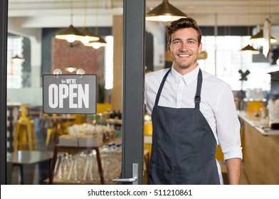 Portrait of smiling owner standing at his restaurant gate with open signboard. Young entrepreneur leaning at the cafeteria door and looking at camera. Chef or waiter standing in front of coffee shop. - Shutterstock ID 511210861