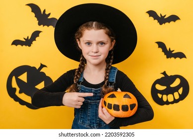 Portrait Of Smiling Pretty Little Kid Girl In Big Hat Point Finger At Orange Funny Pumpkin, Celebrates Halloween Holiday, Posing Isolated Over Yellow Color Background Wall In Studio With Copy Space