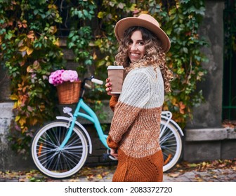 Portrait of smiling pretty woman with curly hair in beige hat holding cup tasty hot coffee or tea on vintage blue bicycle background. Concept of walking on fresh air in centre city. - Powered by Shutterstock