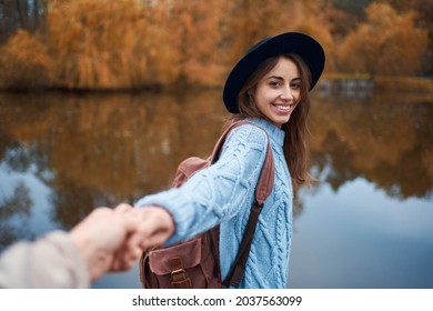 Portrait smiling pretty woman in hat holding hand of her boyfriend while walking by lake at autumn park. Couple enjoying autumn time - Powered by Shutterstock