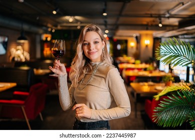 Portrait of smiling pretty woman holding glass of red wine standing in restaurant with luxury interior, looking at camera. Front view of blonde positive female posing at cafe, blurred background - Powered by Shutterstock