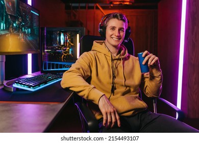 Portrait Of Smiling Pro Gamer Wearing Headset, Looking At Camera, Holding Energy Caffeine Dink, Sitting On Gaming Chair Near Professional Gaming Setup. Cyber Sportsman Playing Shooter Computer Game