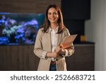 Portrait of smiling professional businesswoman in business casual outfit standing at corporate office with clipboard in her hands and looking at camera. Confident successful employee posing at office.