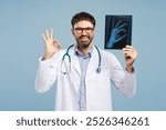 Portrait of smiling professional doctor, attractive man wearing white coat, holding X-ray picture showing ok sign, looking at camera, isolated on blue background. Health care concept