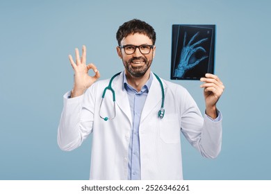 Portrait of smiling professional doctor, attractive man wearing white coat, holding X-ray picture showing ok sign, looking at camera, isolated on blue background. Health care concept - Powered by Shutterstock