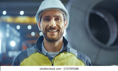Portrait of Smiling Professional Heavy Industry Engineer / Worker Wearing Safety Uniform and Hard Hat. In the Background Unfocused Large Industrial Factory
