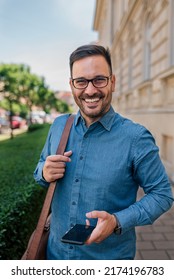 Portrait Of Smiling Professional With Smart Phone On Sidewalk By Plants. Happy Male Executive Commuter Is Carrying Laptop Bag. He Is Wearing Formals In The City.