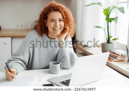 Portrait of smiling redhead curly female student studying at home, doing home work in front of laptop, preparing for exams, sitting at kitchen table with pen in hands, looking at camera