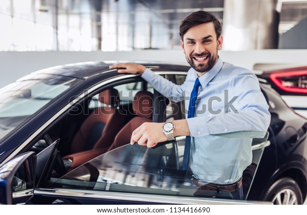 portrait of\
smiling salesman standing at the car with opened door in dealership\
salon and looking at the\
camera