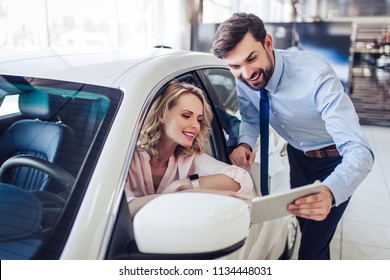 Portrait Of Smiling Salesman Talking With Female Client Sitting In The Car Using Digital Tablet In Dealership Salon