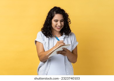 Portrait of smiling satisfied attractive woman with dark wavy hair writing in paper notebook, making notes, schedule or to do list. Indoor studio shot isolated on yellow background. - Powered by Shutterstock