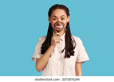 Portrait of smiling satisfied woman with black dreadlocks holding magnifier on her teeth, looking at camera with happy expression, wearing white shirt. Indoor studio shot isolated on blue background. - Powered by Shutterstock