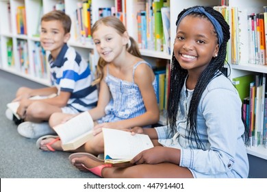 Portrait Of Smiling School Kids Sitting On Floor And Reading Book In Library