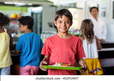 Portrait Of Smiling Schoolboy Holding Food Tray In Canteen Against Classmates