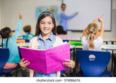 Portrait Of Smiling Schoolgirl Standing With Book In Classroom At Elementary School