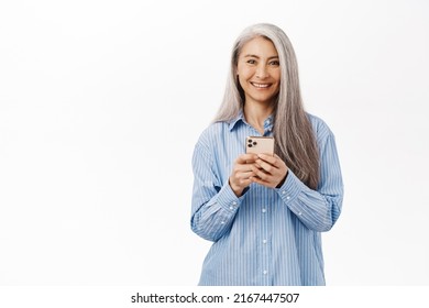 Portrait Of Smiling Senior Asian Woman Using Smartphone. Japanese Old Lady Holding Mobile Phone With Happy Face, Standing Over White Background