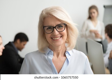 Portrait Of Smiling Senior Businesswoman Wearing Glasses With Businesspeople At Background, Happy Older Team Leader, Female Aged Teacher Professor Or Executive Woman Boss Looking At Camera, Head Shot