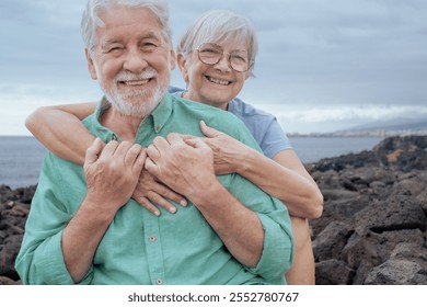 Portrait of smiling senior couple have fun sitting outdoors on the seaside looking at camera laughing. Cloudy sky in background and horizon over water - Powered by Shutterstock