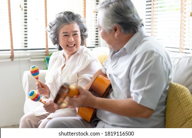 Portrait of smiling senior couple playing guitar singing and her wife holding maracas dancing and sitting on sofa at home. Enjoying lifestyle during retirement life at home.