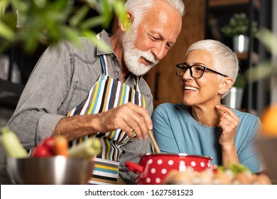 Portrait Of Smiling Senior Couple Standing Close Together, Cuddling Caringly And Laughing, Cooking At Kitchen Table 