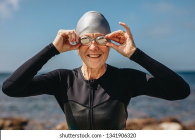 Portrait Of A Smiling Senior Female Swimmer With Sea In The Background. Cheerful Senior Woman In Swim Wear Standing On The Beach Holding Her Swimming Goggles.