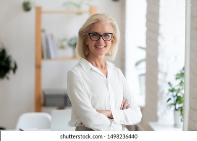 Portrait of smiling senior grey-haired beautiful businesswoman stand with arms crossed posing for picture at workplace, happy confident aged woman worker or company ceo in glasses look at camera