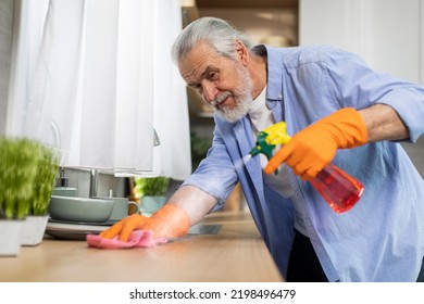 Portrait Of Smiling Senior Man Cleaning Table In Kitchen From Dust, Tidy Elderly Gentleman Using Rag And Spray Detergent For Polishing Surface, Enjoying Doing Domestic Chores, Closeup