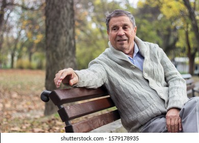 Portrait of a smiling senior man sitting on the bench, in the public park, outdoors. Old man relaxing outdoors and looking away. Portrait of elderly man enjoying retirement