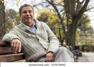 Portrait of a smiling senior man sitting on the bench, in the public park, outdoors. Old man relaxing outdoors and looking at the camera. Portrait of elderly man enjoying retirement
