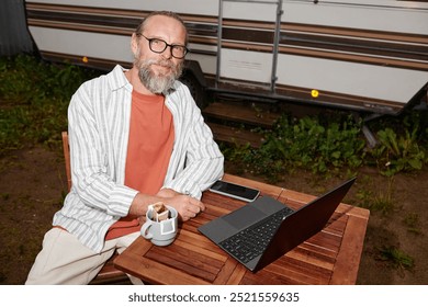 Portrait of smiling senior man using laptop outdoors by van looking at camera, shot with flash, copy space
