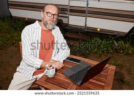 Portrait of smiling senior man using laptop outdoors by van looking at camera, shot with flash, copy space