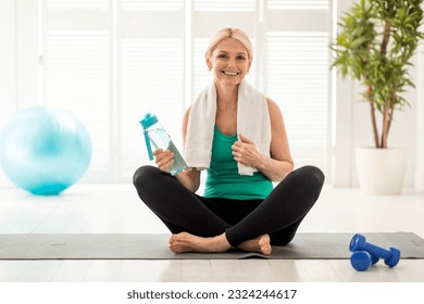 Portrait Of Smiling Senior Woman In Activewear Posing On Yoga Mat At Home, Happy Mature Female Holding Water Bottle And Looking At Camera, Relaxing After Domestic Training, Copy Space - Powered by Shutterstock