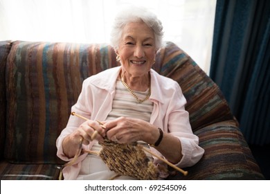 Portrait of smiling senior woman knitting while sitting on sofa against window at retirement home - Powered by Shutterstock