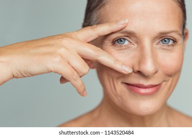 Portrait Of Smiling Senior Woman With Perfect Skin Showing Victory Sign Near Eye On Grey Background.  Closeup Face Of Mature Woman Showing Successful Results After Anti-aging Wrinkle Treatment.