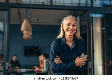 Portrait of smiling senior woman standing in office doorway with her arms crossed. Beautiful female executive at office with people meeting in background. - Powered by Shutterstock