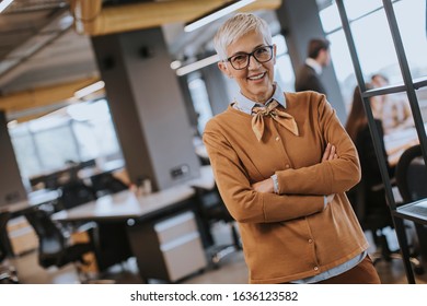 Portrait Of Smiling Senior Woman Standing Outside Meeting Room With Team Discussing Work In Background