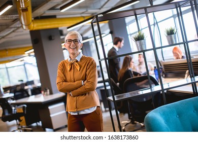 Portrait Of Smiling Senior Woman Standing Outside Meeting Room With Team Discussing Work In Background