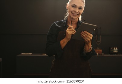 Portrait of smiling senior woman wearing apron holding digital tablet in her jewelry workshop. Smiling mature female goldsmith with tablet pc. - Powered by Shutterstock