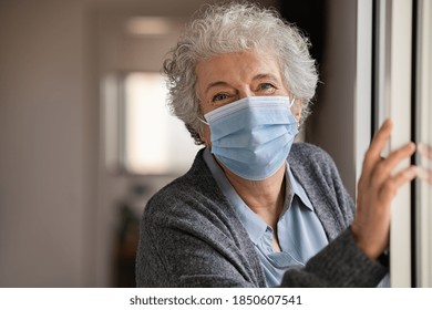 Portrait of smiling senior woman wearing face mask near window. Old woman wearing surgical mask and looking at camera during the lockdown. Happy elderly lady during the covid-19 pandemic stay at home. - Powered by Shutterstock