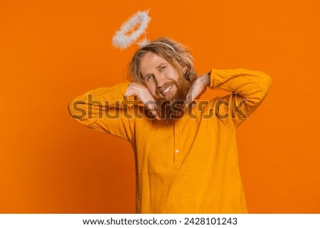 Portrait of smiling shy angelic Caucasian man with angel halo nimb over head flirting, looking at camera, positive emotions, celebrating holidays. Redhead young guy isolated on orange background