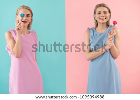 Portrait of smiling sisters holding sweet lollipops. One is covering eye with gum. Isolated on blue and pink background
