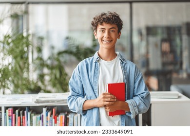 Portrait of smiling smart curly haired teenage boy holding book looking at camera. Back to school, Education concept - Shutterstock ID 2333649091