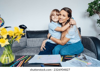 Portrait smiling son hugging his happy mother during home family weekend Mother   son drawing and color pencils   markers in the living room  children creativity concept  Selective focus 