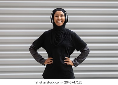 Portrait Of Smiling Sporty Muslim Woman In Hijab And Wireless Headphones Posing Near Urban Wall While Training Outdoors, Fit Athletic Islamic Lady In Modest Sportswear Smiling At Camera, Copy Space - Powered by Shutterstock