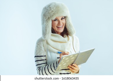 Portrait Of Smiling Stylish Middle Age Woman In White Striped Sweater, Scarf And Ear Flaps Hat With Pen Writing In Notebook Isolated On Winter Light Blue Background.